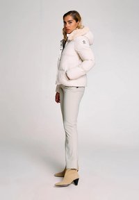 Donna in pantaloni beige, stivali col tacco color sabbia e un giubbotto bianco imbottito con cappuccio foderato di pelliccia, in piedi su uno sfondo neutro.