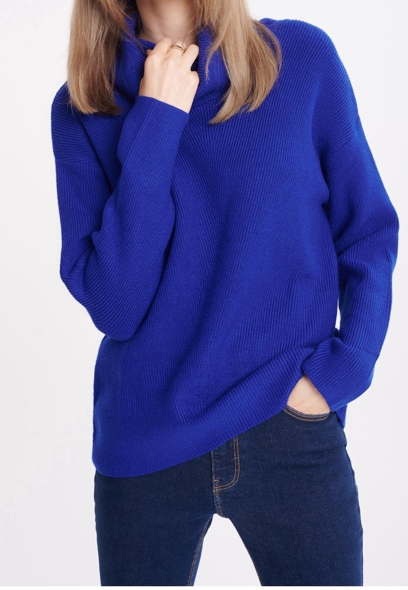Pull bleu en tricot avec un col bénitier, texture côtelée et coupe ample. Associé à un jean en denim foncé, mettant en valeur un style décontracté.