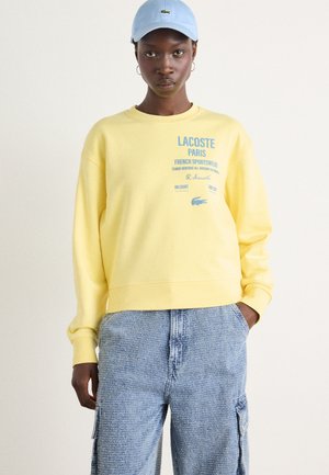Felpa gialla corta con testo e logo stampati in blu, abbinata a pantaloni cargo leggeri blu chiaro con tasche laterali.