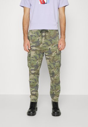 Homme portant un pantalon cargo camouflage avec des bas élastiques, des bottes noires à lacets et un t-shirt violet clair avec un imprimé graphique.