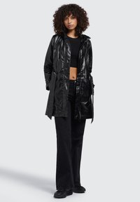 Trench coat impermeabile nero con una finitura lucida, dotato di zip sul davanti, cappuccio regolabile e tasche laterali, abbinato a jeans e scarpe neri.