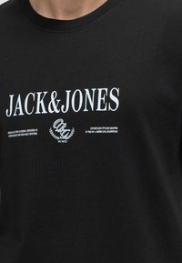 Μαύρο μπλουζάκι με λευκό κείμενο "JACK & JONES" στο κέντρο και μικρότερες λευκές τυπωμένες φράσεις κάτω από το όνομα της μάρκας.