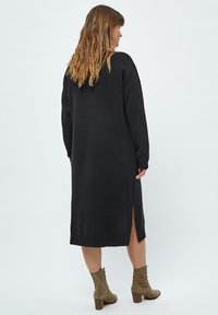 Schwarzes Langarmkleid aus weichem Stoff, mit geradem Schnitt, seitlichen Schlitzen und einem gerippten Saum. Kombiniert mit beigen Stiefeletten.