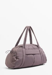 Puhán párnázott duffel táska lágy, tompított lila színben, dupla fogantyúval, elülső cipzáras zsebbel és lekerekített formával, texturált felülettel.