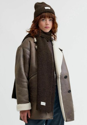 Brown leather jacket with white shearling lining and black buttons, paired with a dark brown knitted scarf and beanie.