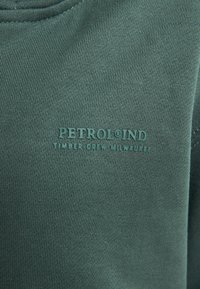 Groene sweatshirt van zacht materiaal met een gestructureerde afwerking. De voorkant heeft reliëfletters: "PETROL®IND TIMBER CREW MILWAUKEE."