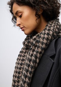Scarf in motivo houndstooth marrone e nero realizzato in tessuto morbido, avvolto attorno al collo, con una texture liscia e una caduta rilassata.