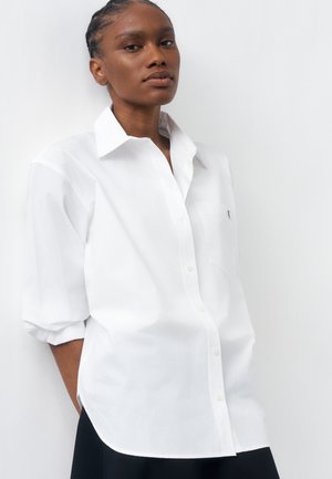 Donna con capelli intrecciati che indossa una camicia bianca ampia con bottoni e maniche arrotolate fino ai gomiti, in piedi davanti a uno sfondo bianco semplice.