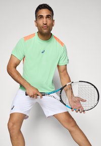 Jucător de tenis masculin îmbrăcat în echipament sportiv verde mentă și alb, ținând o rachetă, pregătit să returneze o minge pe un fundal simplu.