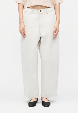 Personne portant un pantalon blanc à taille haute, ample avec jambes larges, équipé de boutons et d'une fermeture éclair, associé à des ballerines noires et un haut court beige.
