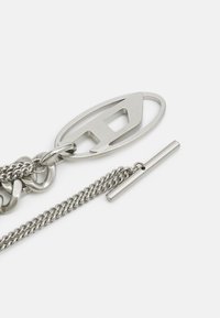 Accesorio de cadena de metal plateado que presenta un charm de logotipo ovalado y un cierre cilíndrico, con un acabado texturizado y varios tamaños de eslabones.