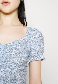 Blusa floral en azul claro, presenta un escote cuadrado, mangas abullonadas y textura plisada con pequeños patrones de flores.
