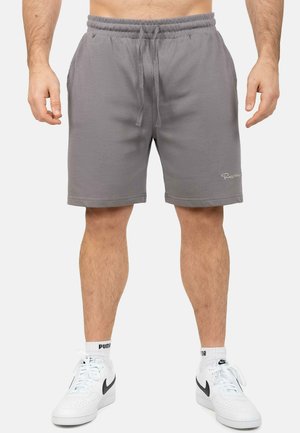 Mann trägt graue Shorts mit Kordelzug, weiße Nike-Sneaker und weiße Puma-Socken, steht mit entspannten Beinen und Armen vor einem einfarbigen Hintergrund.