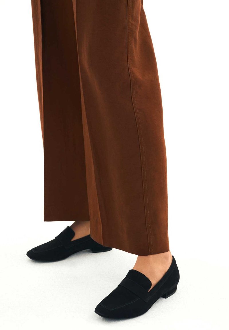 Jambes portant un pantalon large marron et des mocassins en suède noir sur un fond blanc.