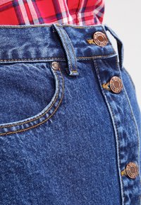 Mini falda de mezclilla en azul medio con un diseño de cintura alta. Presenta botones de color cobre, costuras detalladas y bolsillos laterales. Textura suave.