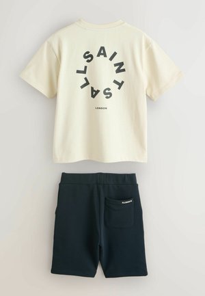 Tricou crem cu mâneci scurte, imprimeu circular negru și text, asortat cu șorturi bleu închis cu buzunar lateral și logo.