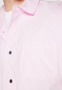 Camisa de botones rosa claro hecha de algodón. Cuenta con un bolsillo en el pecho con el logo bordado "obey" y botones negros.