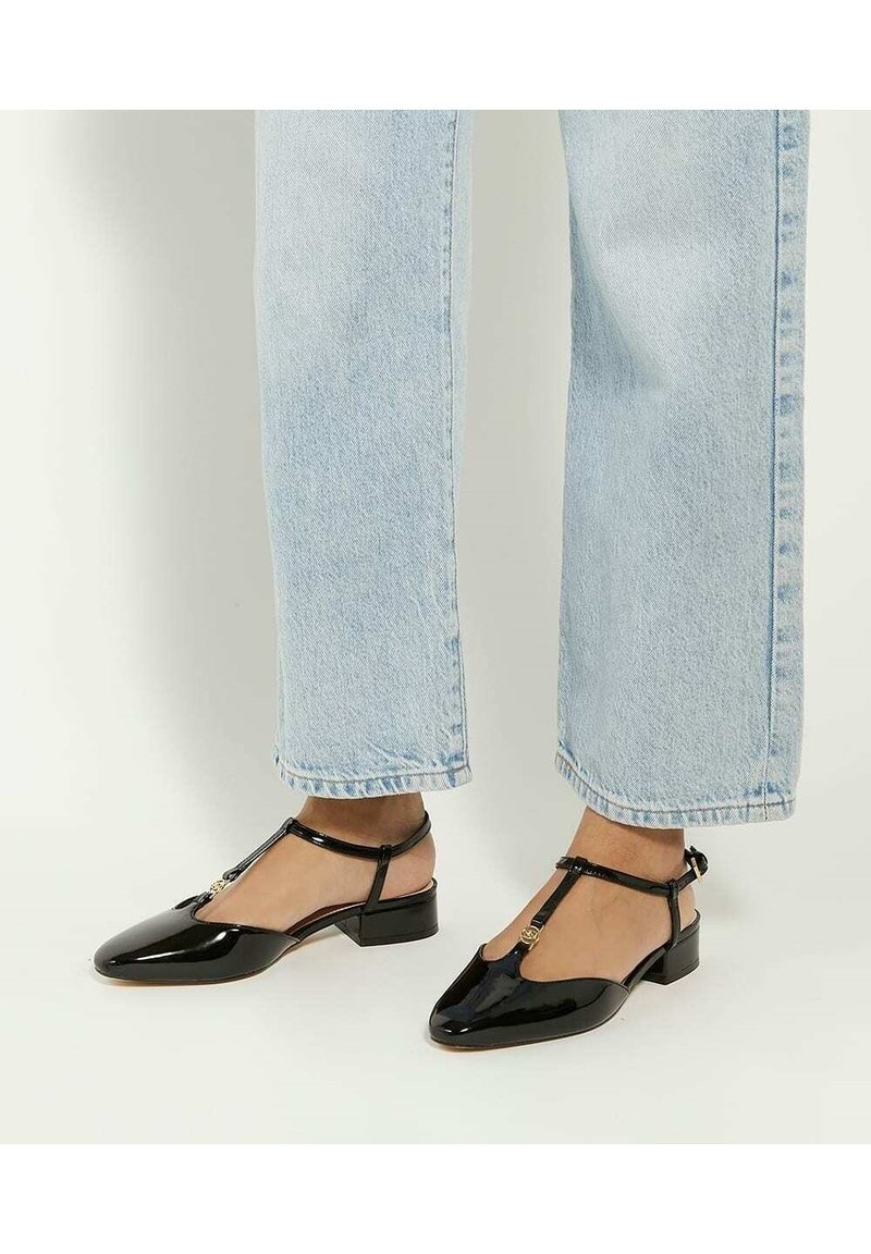Personne portant un jean bleu clair et des chaussures à talons bas en cuir verni noir avec bride en T, debout sur un sol clair.
