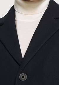 Schwarzer Wollmantel mit Reverskragen und Ein-Knopf-Verschluss, getragen über einem hellbeigen, gerippten Rollkragenpullover. Glatte Textur.