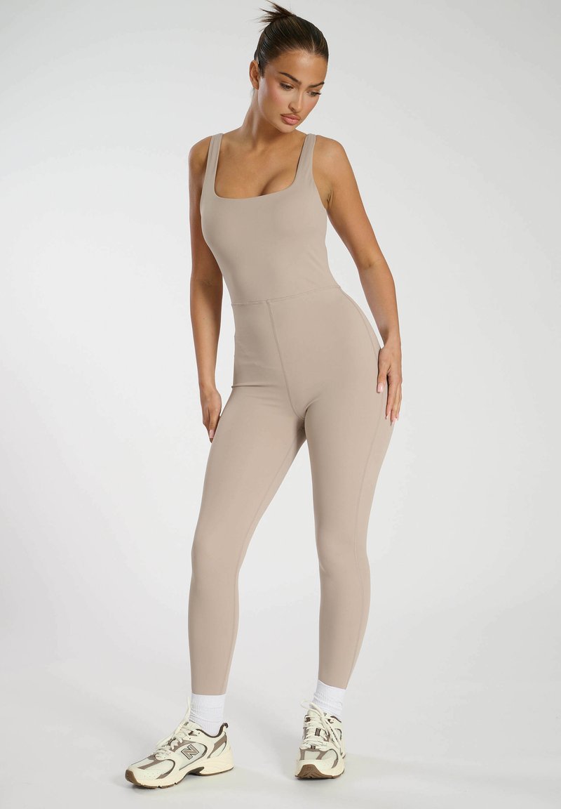 Tight beige jumpsuit med fyrkantig halsringning, tillverkad av jämn tyg. Kombinerad med vita strumpor och krämfärgade sneakers.