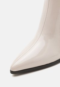 Bottine brillante ivoire à bout pointu avec couture visible et semelle noire, présentée sur fond blanc.