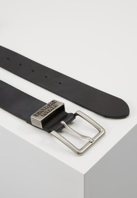 Black leather belt with a silver metal buckle, featuring engraved "ARAMANI EXCHANGE." Smooth texture with evenly spaced holes for adjustment.