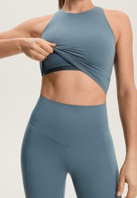Sportovní podprsenka a legíny z tlumeně modré látky; přiléhavý design s hladkou texturou a atletickým střihem. Poskytuje odhalení břicha.