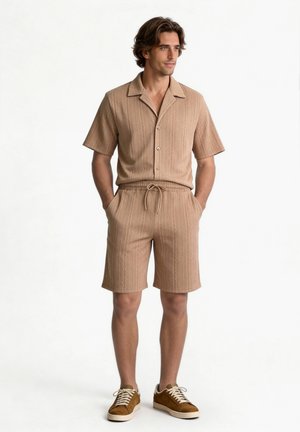 Mann steht mit Händen in den Taschen, trägt ein beiges, strukturiertes kurzärmliges Hemd mit Knöpfen und passende Shorts sowie braune Sneakers.