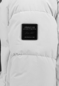 Weißer Pufferjacke mit gestepptem Design, glatter Textur und einem rechteckigen schwarzen Logoschild auf dem oberen Ärmel mit geprägtem Text.