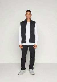 Melna sastāvdaļa bez piedurknēm virs balta garajiem piedurkņu krekla, melnām biksēm un pelēkiem čības. Iekļauj rāvējslēdzēja priekšpusi un logo uz krūts.