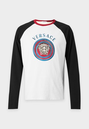 Jeune homme portant un t-shirt Versace blanc et noir, un pantalon large gris à fines rayures, des baskets et un sac d'épaule noir sur fond blanc uni.