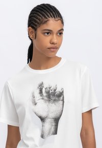 T-shirt en coton blanc avec un graphique d'un poing levé en noir, placé au centre sur le devant. Col rond et manches courtes.