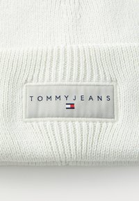 Bonnet en tricot blanc avec un motif texturé. Le patch logo centré indique "TOMMYJEANS" avec des accents bleu, rouge et blanc.