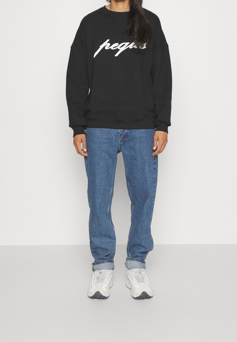 Personne portant un sweat-shirt noir avec le texte "peque", un jean bleu avec revers et des baskets blanches, debout devant un fond clair uni.