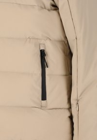 Beige vadderad jacka med en dragkedjeficka med svart kant och subtila sömdetaljer, mjuk textur och en elegant, strömlinjeformad design.