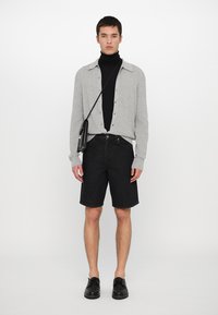 Pelēks ribots krekliņš ar pogām virs melnā dzelsakņa, kombinēts ar melniem džinsu šortiem. Melni apavi un maza crossbody soma. Vienkāršs fons.