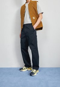 Chaqueta sin mangas marrón sobre una camiseta gris, pantalones cargo azul marino y zapatillas negras con detalles amarillos. Fondo azul plano.