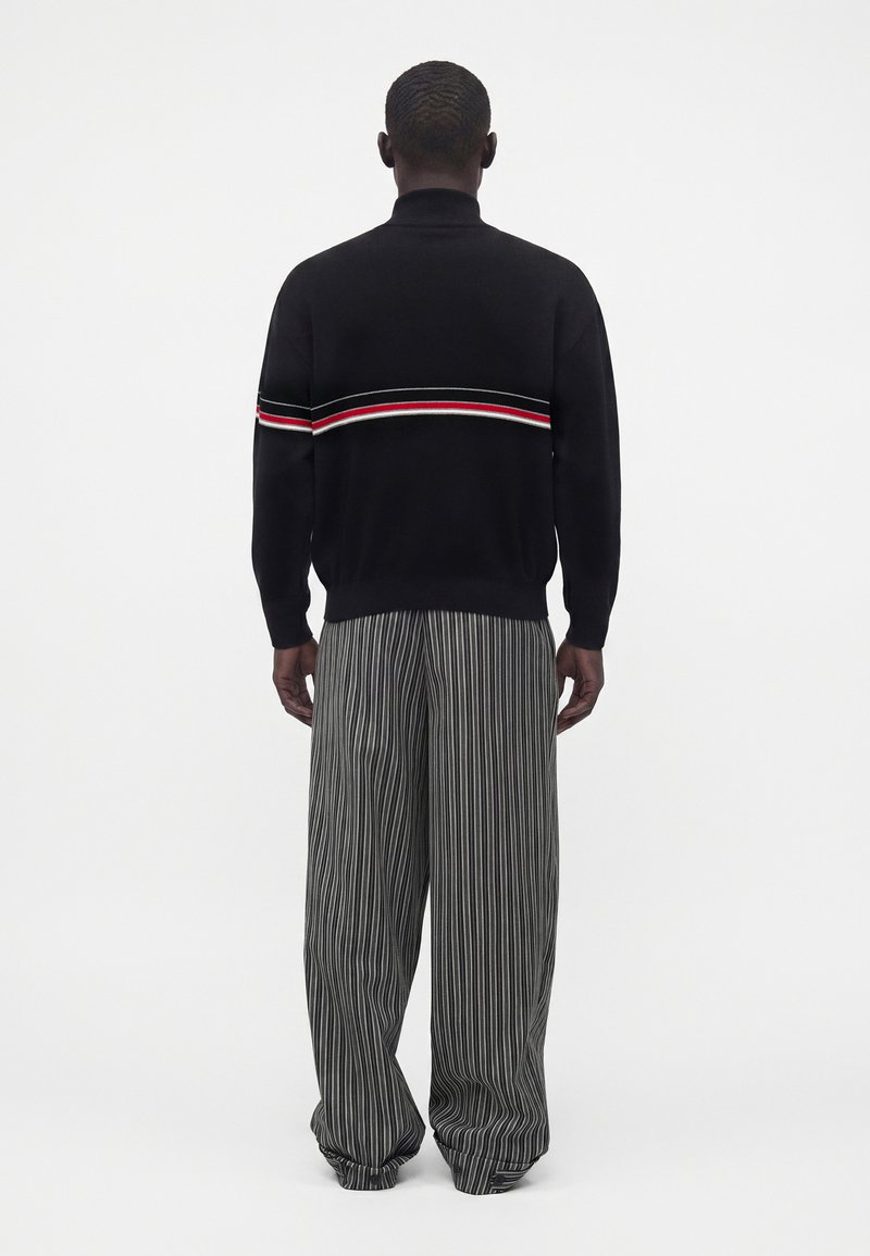 Homme debout de dos, vêtu d’un pull noir avec une bande rouge et blanche et d’un pantalon gris large à fines rayures.