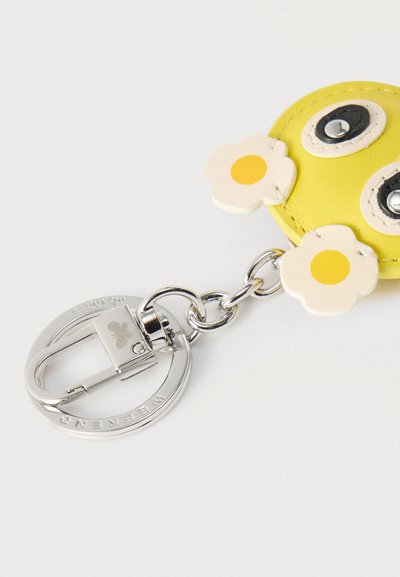 Κίτρινο δερμάτινο μπρελόκ σε στρογγυλό σχήμα, με χαμογελαστό πρόσωπο, μαύρα και λευκά μάτια, και δύο κρεμ λουλούδια με κίτρινο κέντρο. Ασημένια μεταλλικά στοιχεία.