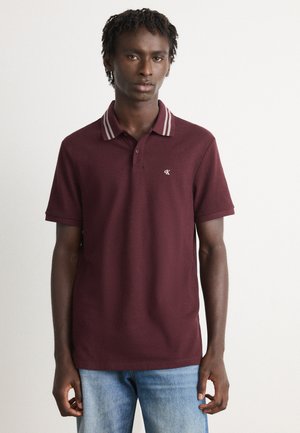 Young man wearing a maroon polo shirt with striped collar and blue jeans, standing against a plain light background.