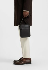 Svart rektangulär crossbody-väska med en framficka med dragkedja, slät textur och diskret branding. Läderrem fäst för bärande.