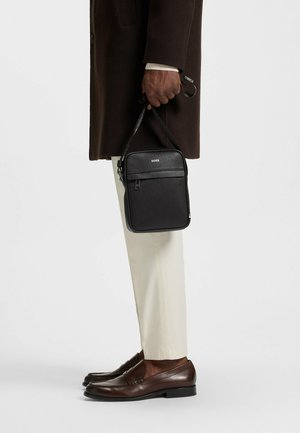 Zwarte rechthoekige crossbody tas met een voorzak met rits, een gladde textuur en subtiele branding. Leren hengsel bevestigd voor het dragen.
