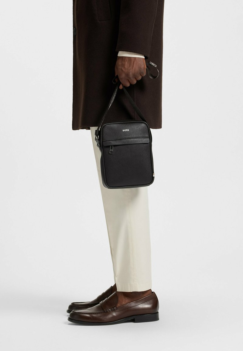 Svart rektangulär crossbody-väska med en framficka med dragkedja, slät textur och diskret branding. Läderrem fäst för bärande.