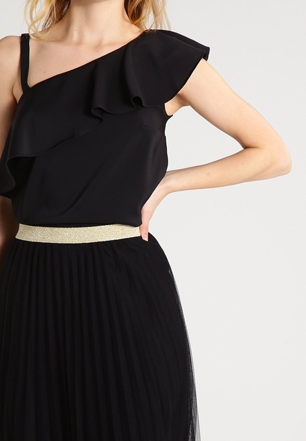 Femme portant un haut noir à une épaule avec volants et une jupe plissée noire avec une ceinture dorée, debout, main sur la hanche.
