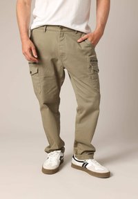 Beige cargobroek met meerdere zakdetails, sluiting met een knoop aan de voorkant, rechte pijpen en een gladde textuur, gecombineerd met witte sneakers.