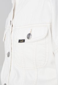 Gros plan sur la poche d'une veste en denim blanche avec rabat, boutons argentés de la marque Lee et petite étiquette noire Lee.
