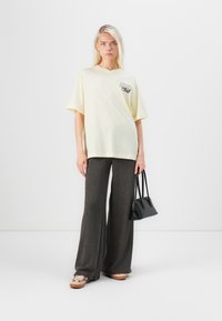 Žluté oversized tričko s grafikou na hrudi, kombinované se šedými ribbed širokými kalhotami a béžovými slip-on botami. Modelka drží černou kabelku.