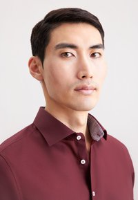 Camicia button-up bordeaux con un colletto testurizzato e decorato. Presenta un tessuto liscio e bottoni bianchi al collo e ai polsini.