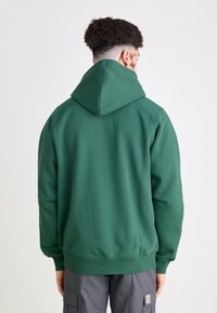 Grüne Kapuzenpullover mit glatter Textur, gerippten Bündchen und Saum sowie gefütterter Kapuze. Trägt eine Person, die von hinten gezeigt wird.