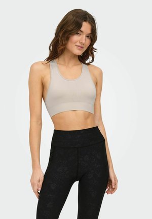 Young woman wearing a beige sports bra and high-waisted black leggings with subtle floral pattern, standing against a light background.